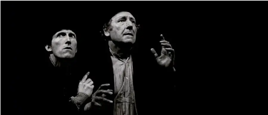 Fotografía en blanco y negro de dos actores en escena (representando a Vladimir y Estragón en Esperando a Godot), con fondo oscuro y gestos teatrales.