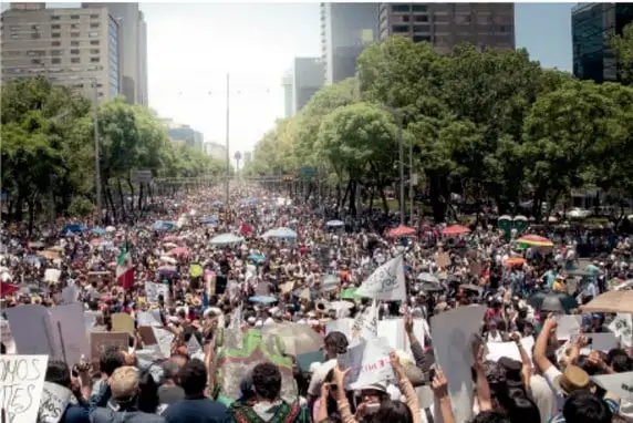 Fotografía de una gran marcha ciudadana en una avenida amplia de una ciudad, con multitud de personas portando pancartas y carteles, entre edificios altos y árboles; representa la exigencia colectiva de derechos.