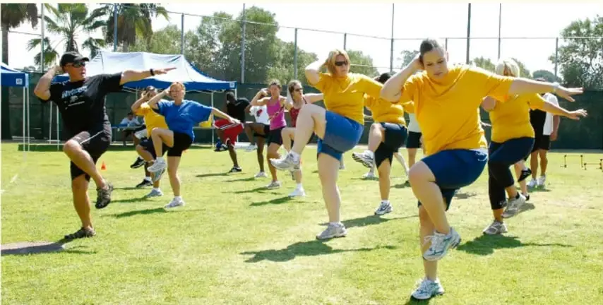 Grupo de personas al aire libre en una cancha de césped realizando ejercicios de calentamiento con el brazo y la rodilla levantados; visten playeras amarillas y azules.