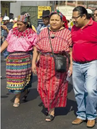 Fotografía de un grupo de personas caminando en una calle; destacan mujeres con trajes tradicionales mayas: huipiles bordados de colores y faldas tejidas rojas con diseños geométricos.