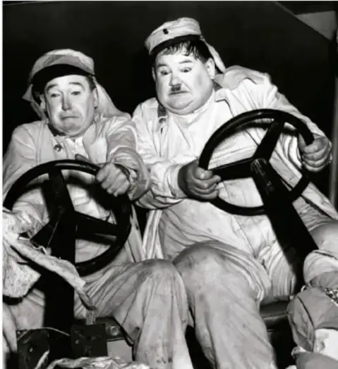 Fotograma en blanco y negro del dúo cómico Laurel y Hardy (El Flaco Stan Laurel a la izquierda y El Gordo Oliver Hardy a la derecha) sentados frente a volantes, ambos con uniforme.