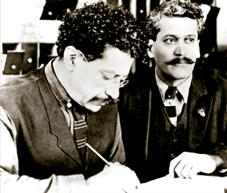 Fotografía histórica en blanco y negro de Ricardo y Enrique Flores Magón (ca. 1908). A la izquierda, Ricardo Flores Magón con cabello rizado, lentes y bigote, inclinado escribiendo con un lápiz sobre un documento; a la derecha, Enrique Flores Magón con cabello ondulado y bigote, mirando hacia el frente, con moño y chaleco. Procedencia: Biblioteca del Congreso de Estados Unidos.