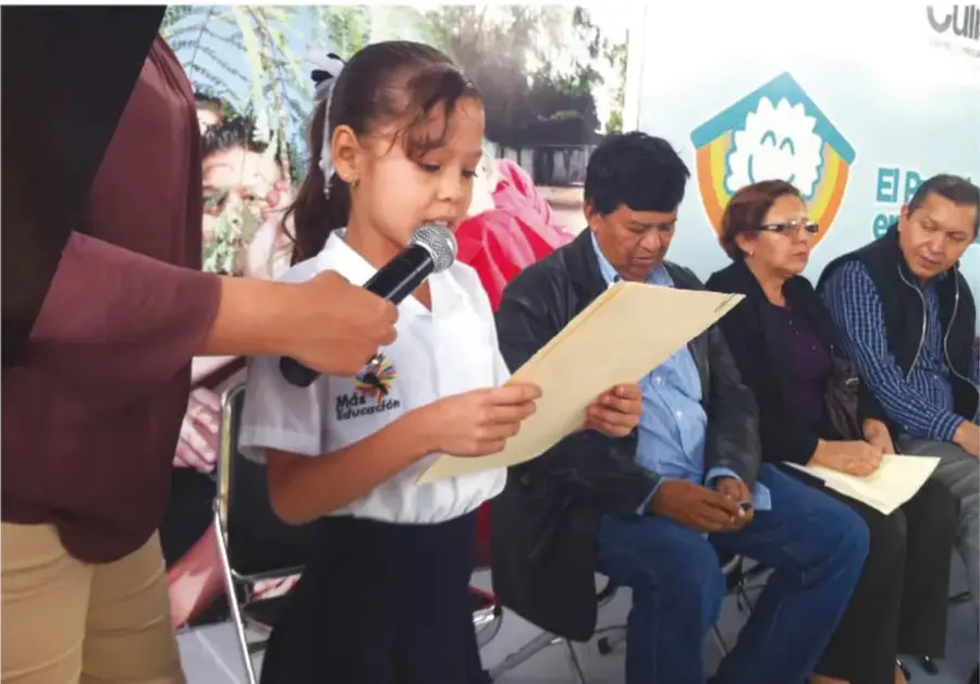 Fotografía de una niña de primaria leyendo un discurso frente a un micrófono, acompañada por adultos sentados que la escuchan; al fondo hay un cartel con el logotipo de un programa escolar.