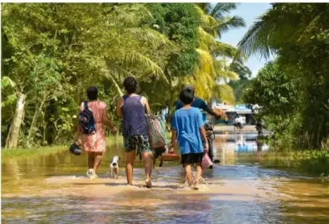 Fotografía de tres personas caminando por una calle inundada en una zona con vegetación tropical, ilustrando las consecuencias de los fenómenos climáticos extremos.