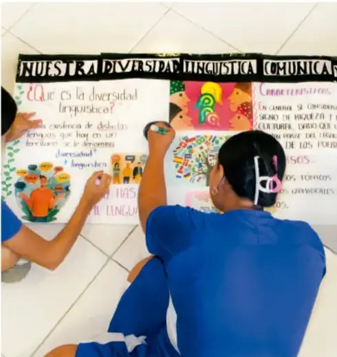 Fotografía de estudiantes elaborando un cartel titulado "Nuestra diversidad lingüística comunica", con la pregunta "¿Qué es la diversidad lingüística?" y dibujos de personas; una estudiante escribe con plumón en el cartel.