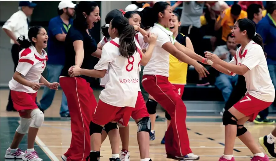 Fotografía de un equipo escolar de voleibol (uniformes blancos con detalles rojos, número 8 visible, texto 'NUEVO LEÓN') celebrando con entusiasmo junto con su entrenadora tras un punto, en una cancha techada con público en las gradas.