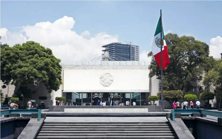 Fotografía a color de la fachada del Museo Nacional de Antropología, con una bandera de México al frente, escalinatas, árboles y visitantes.
