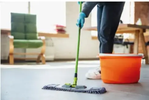 Persona limpiando el piso con un trapeador y un balde naranja en un interior ordenado.