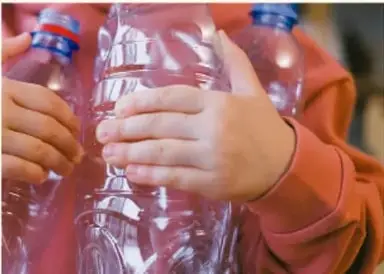 Fotografía de las manos de un estudiante sosteniendo botellas de plástico transparente con tapón azul, material para armar los bolos del boliche.