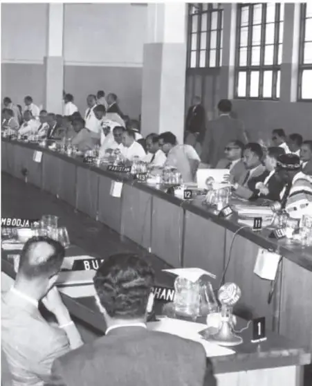 Fotografía en blanco y negro de una sesión de la Conferencia de Bandung, con delegados sentados en una mesa larga y carteles con nombres de países como MBOOJA y RU.