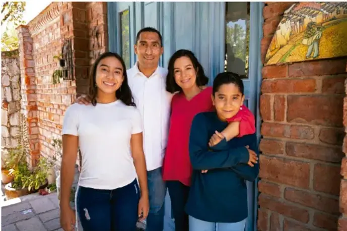 Fotografía de una familia de cuatro personas (dos adultos y dos jóvenes) sonriendo frente a la entrada de una casa de ladrillo, ilustrando el ámbito familiar.
