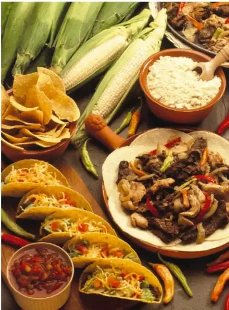 Mesa con platillos mexicanos: tacos, un plato con guiso, totopos o papas fritas de maíz, elotes, un cuenco con arroz y salsas roja y verde.
