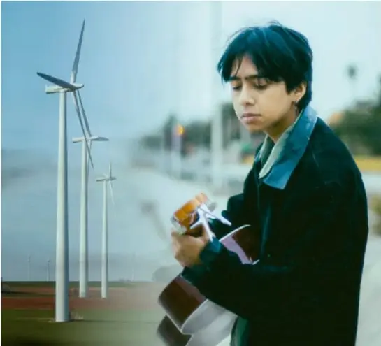 Fotografía de un joven que camina por una zona con aerogeneradores (molinos de viento) al fondo, tocando un instrumento de viento, lo que representa la energía eólica y la expresión artística del proyecto.