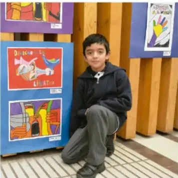Niño arrodillado junto a una exposición de carteles y dibujos sobre migración, colocados sobre paneles de madera.