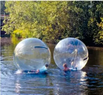 Fotografía de dos esferas acuáticas transparentes con personas dentro, flotando sobre un río rodeado de árboles.