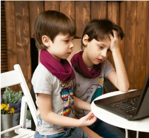 Dos niños pequeños sentados frente a una computadora portátil sobre una mesa redonda blanca, uno de ellos con la mano en la cabeza, en un ambiente con pared de madera.