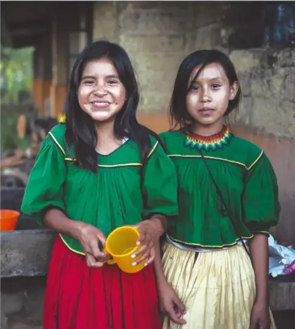 Fotografía de dos niñas indígenas sonrientes con blusas verdes de manga abullonada y faldas (una roja y otra beige con detalles), una sostiene un jarro amarillo; se aprecia un ambiente rural tradicional.