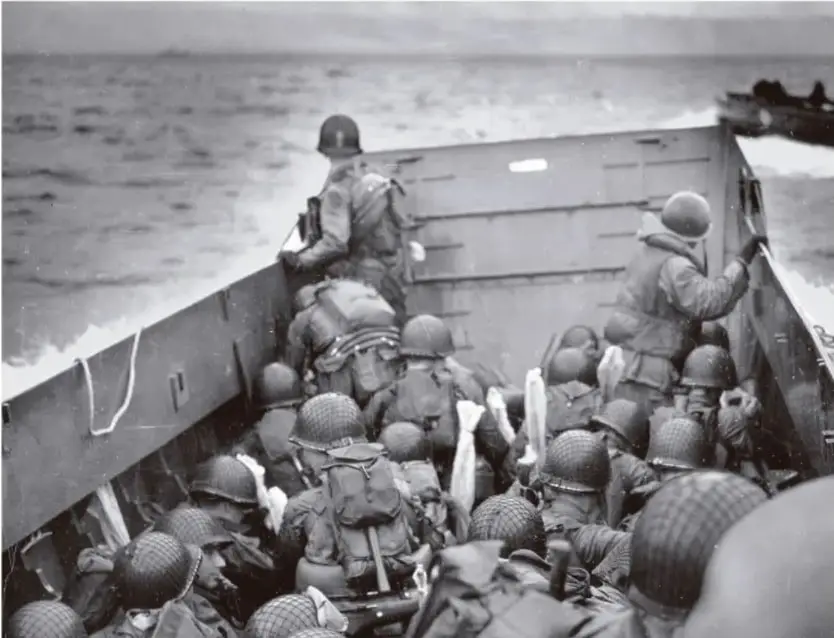 Fotografía en blanco y negro de soldados con casco descendiendo de una lancha de desembarco en el mar durante la Segunda Guerra Mundial.