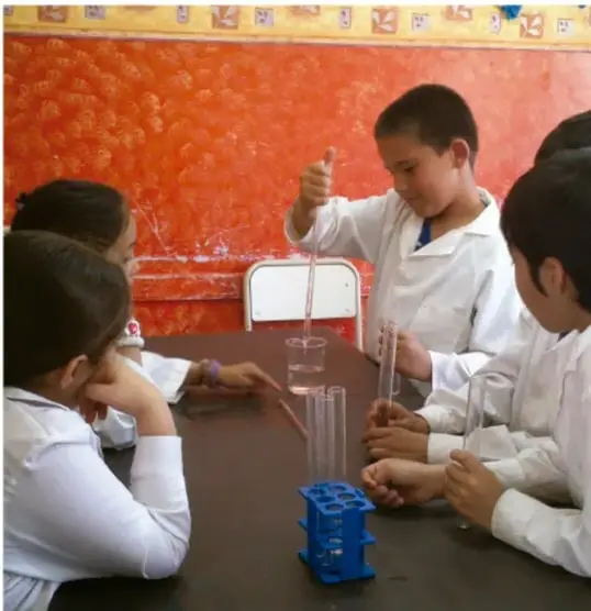 Fotografía de alumnos con batas blancas realizando un experimento de laboratorio con matraces, probetas y portamuestras azul, en un salón con pared de ladrillo rojo, ilustrando la práctica científica escolar.