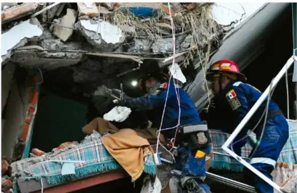 Rescatistas con cascos y uniformes azules trabajando entre escombros de un edificio colapsado; uno de ellos sostiene a una persona.