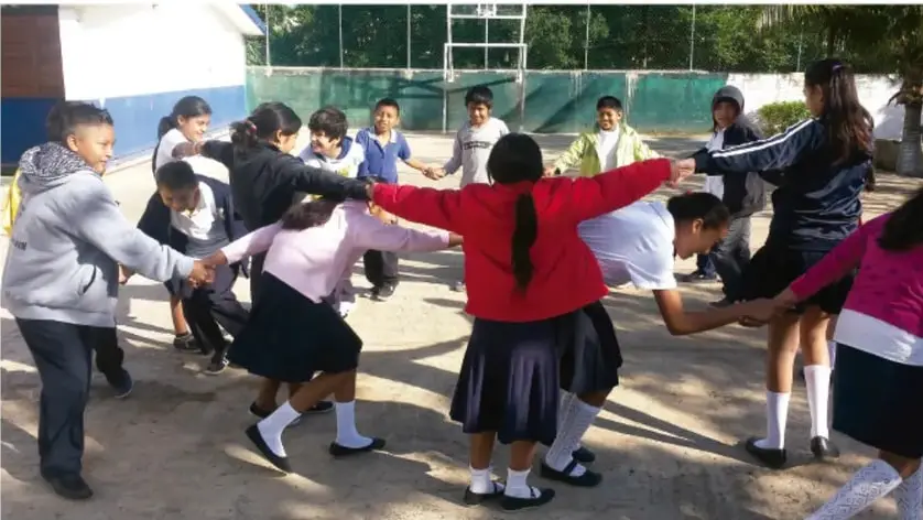 Fotografía de un grupo de estudiantes de secundaria con uniforme formando un círculo tomados de las manos, jugando en el patio de una escuela.