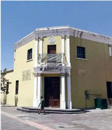 Fotografía de la fachada de un edificio histórico de dos niveles, pintado de color ocre, con columnas, puerta en arco y un pequeño balcón con reja; una persona con ropa verde camina frente a él. El letrero del edificio no es legible con certeza.