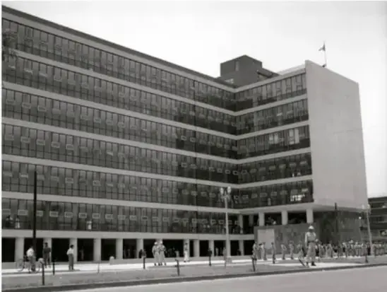 Fotografía en blanco y negro de un edificio moderno de varios pisos con fachada de ventanas, probablemente una dependencia gubernamental de la época.