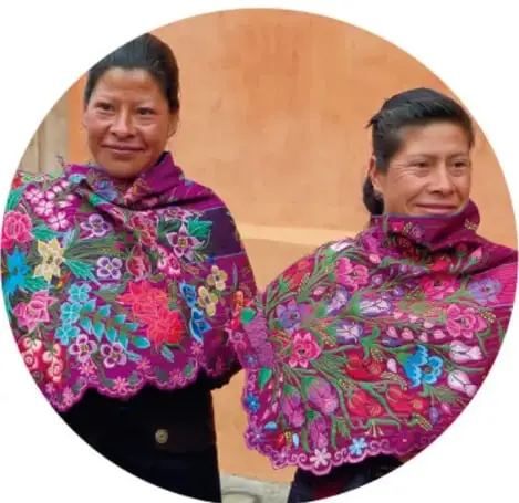 Dos mujeres mayas sonrientes vistiendo coloridos huipiles y chales bordados de flores.