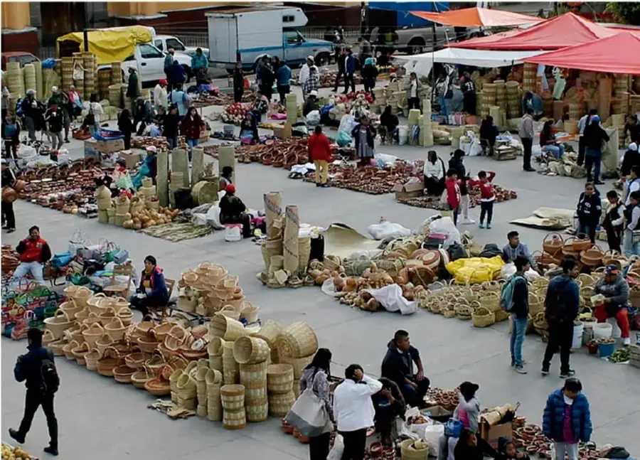 Fotografía de un mercado o tianguis al aire libre con puestos de canastas de mimbre, artesanías y productos locales; se observan vendedores y compradores, algunos con bolsas de tela.