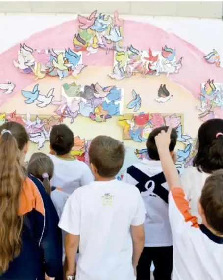 Fotografía de niñas y niños de espaldas observando un mural colectivo colorido con palomas de colores y figuras pintadas; representa la socialización de los productos de la campaña por la paz.