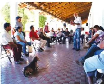 Otra reunión comunitaria en un espacio techado, personas sentadas en sillas, una de pie hablando, un perro presente.