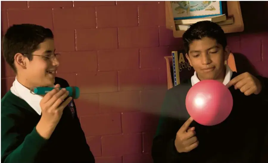 Dos estudiantes en una sala con muralla de ladrillos. El de la izquierda, con lentes y uniforme verde, sostiene una linterna verde y la dirige hacia una pelota rosada que sostiene su compañero; se observa un haz de luz sobre la pelota. Detrás hay un estante con libros.