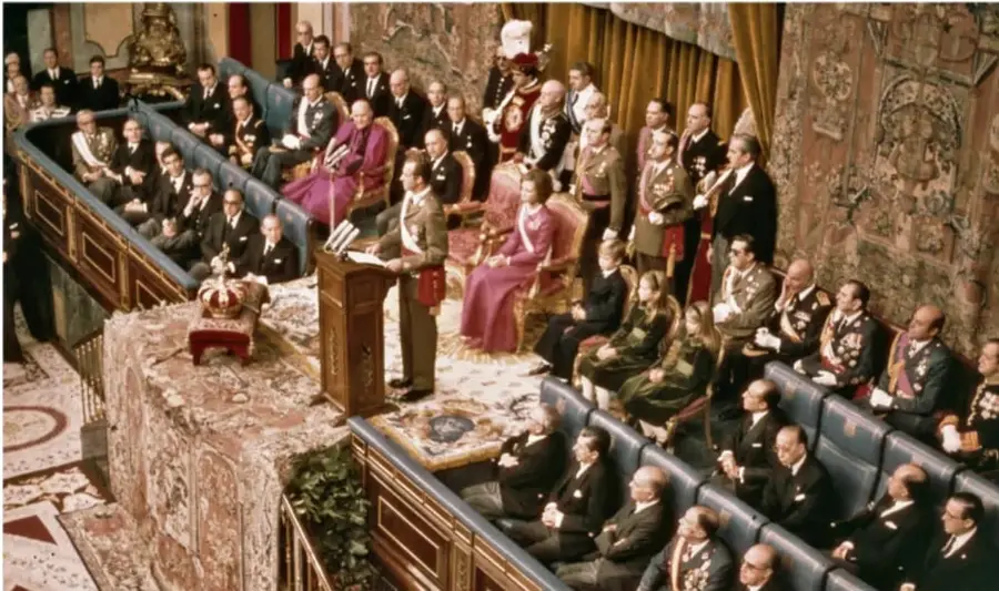 Fotografía de una sesión solemne en un recinto con decoración suntuosa. Se observa a Juan Carlos I de pie en un atril, rodeado de autoridades civiles y militares, algunos sentados en bancos y otros de pie. La escena parece ser un acto de proclamación o juramento.
