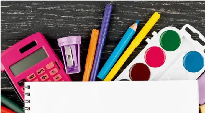 Fotografía de materiales escolares sobre una superficie de madera oscura: calculadora rosa, sacapuntas morado, colores (naranja, morado, azul, amarillo), paleta de acuarelas (verde, rosa, azul) y un cuaderno de espiral abierto en primer plano.