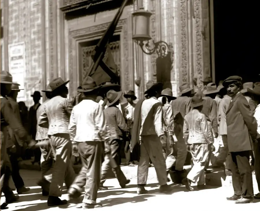 Fotografía histórica en sepia de un grupo de trabajadores con sombreros y ropa de trabajo entrando al Palacio de los Azulejos, sede de la Casa del Obrero Mundial, en 1915; se distingue una bandera y la fachada del edificio.