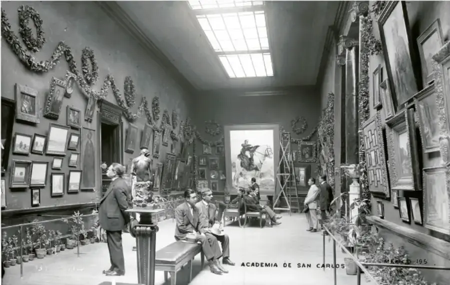 Fotografía en blanco y negro de una sala de museo con cuadros en las paredes, visitantes y una escalera; al pie se lee 'ACADEMIA DE SAN CARLOS'.