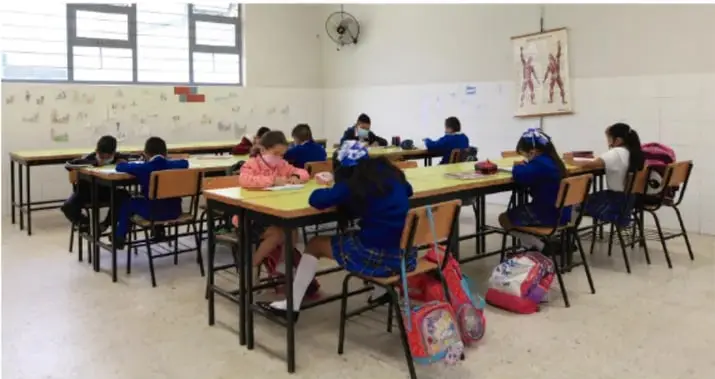 Fotografía de un aula de primaria con estudiantes sentados en mesas, trabajando en grupo. Al fondo hay una ventana y un ventilador de pared.