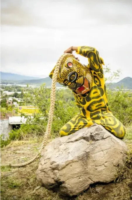 Persona vestida de tigre (traje amarillo con rayos negros y máscara de jaguar) en cuclillas sobre una enorme roca, en un paisaje con montañas y pueblo al fondo; sostiene una cuerda.