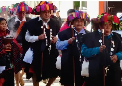 Grupo de personas con vestimenta tradicional y tocados coloridos portando bastones durante la procesión de cambio de autoridades.