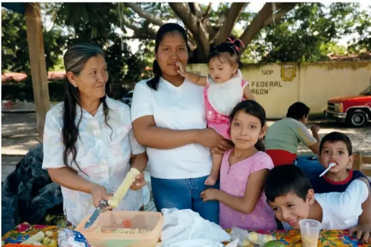 Grupo familiar reunido alrededor de una mesa con alimentos: una señora mayor, una mujer joven con un bebé, una niña y dos niños; al fondo un muro con la leyenda 'FEDERAL' y un vehículo rojo.
