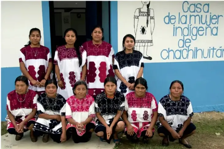 Fotografía de un grupo de mujeres indígenas frente a un mural que dice 'Casa de la Mujer Indígena de Chachihuitán', vestidas con trajes tradicionales bordados.