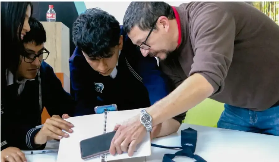 Fotografía de un docente acompañando a tres estudiantes alrededor de una mesa; revisan juntos un cuaderno y un teléfono celular, en un ambiente escolar.