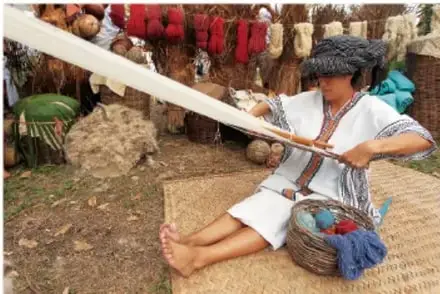 Fotografía de una mujer indígena sentada en el suelo, hilando fibra con un malacate. Viste ropa tradicional y tiene una canasta con ovillos de hilo de colores.