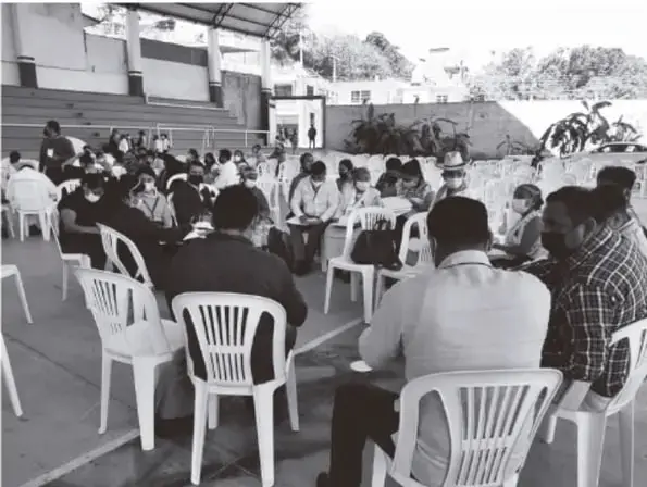 Fotografía en blanco y negro de una asamblea o reunión comunitaria: personas adultas sentadas en sillas de plástico blancas dispuestas en círculo en un patio o techado abierto, dialogando; evoca la asamblea comunitaria como forma de organización y aprendizaje colectivo.