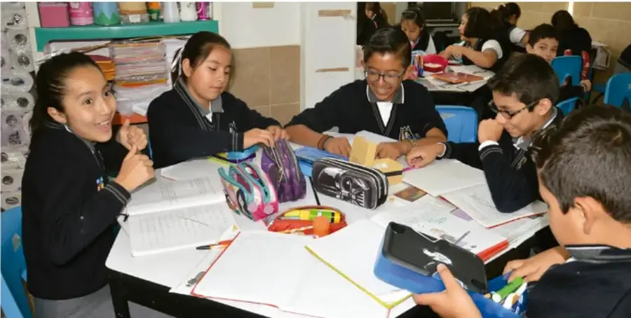 Fotografía de estudiantes de primaria sentados alrededor de una mesa, trabajando en equipo con cuadernos, colores y una tableta.