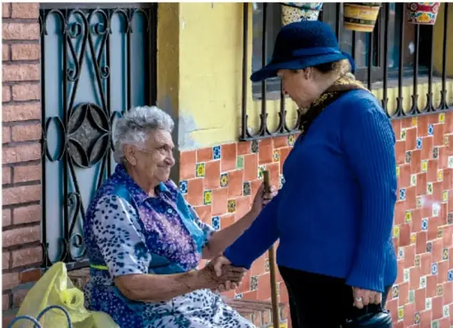 Fotografía de dos mujeres mayores conversando y tomadas de la mano sentadas frente a una pared con azulejos, representando la convivencia y el apoyo mutuo en la comunidad.