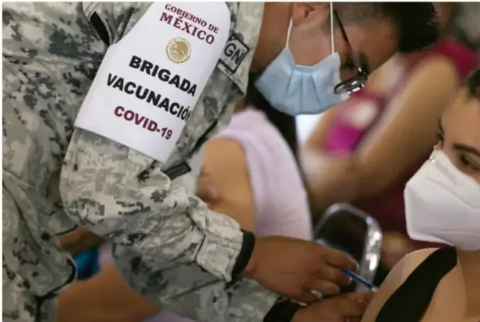 Personal militar con brazalete 'Brigada Vacunación COVID-19, Gobierno de México' aplicando una vacuna en el brazo de una persona con cubrebocas durante la campaña de vacunación.