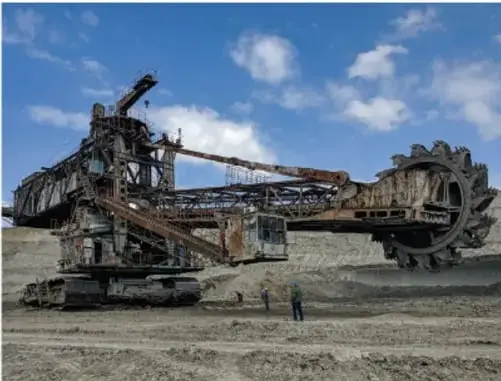 Excavadora de rueda de cuchillas (bucket-wheel excavator) de gran escala operando en una mina a cielo abierto, con personal de mina de pie al pie de la maquina, lo que muestra su enorme tamaño.