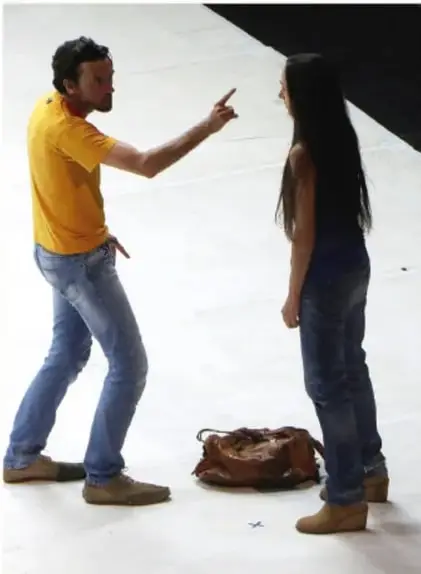 Fotografía de un hombre de camiseta amarilla que señala con el dedo a una mujer joven; ella está de espaldas con una mochila en el suelo. Representa una situación de violencia verbal o psicológica.