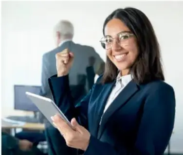 Fotografía de una mujer joven con lentes, vestida formalmente, sosteniendo una tableta y sonriendo; al fondo se ve un hombre mayor de espaldas.